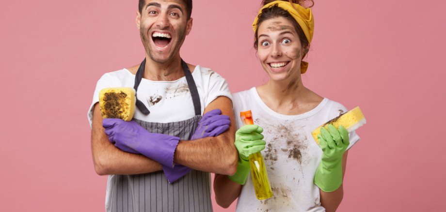 Excited man in casual clothes keeping hands crossed holding dirty sponge rejoicing his work. Smiling woman wearing yellow headband and white T-shirt holding detergent and sponge washing windows