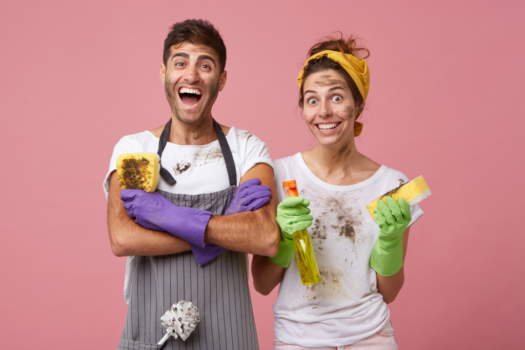 Excited man in casual clothes keeping hands crossed holding dirty sponge rejoicing his work. Smiling woman wearing yellow headband and white T-shirt holding detergent and sponge washing windows